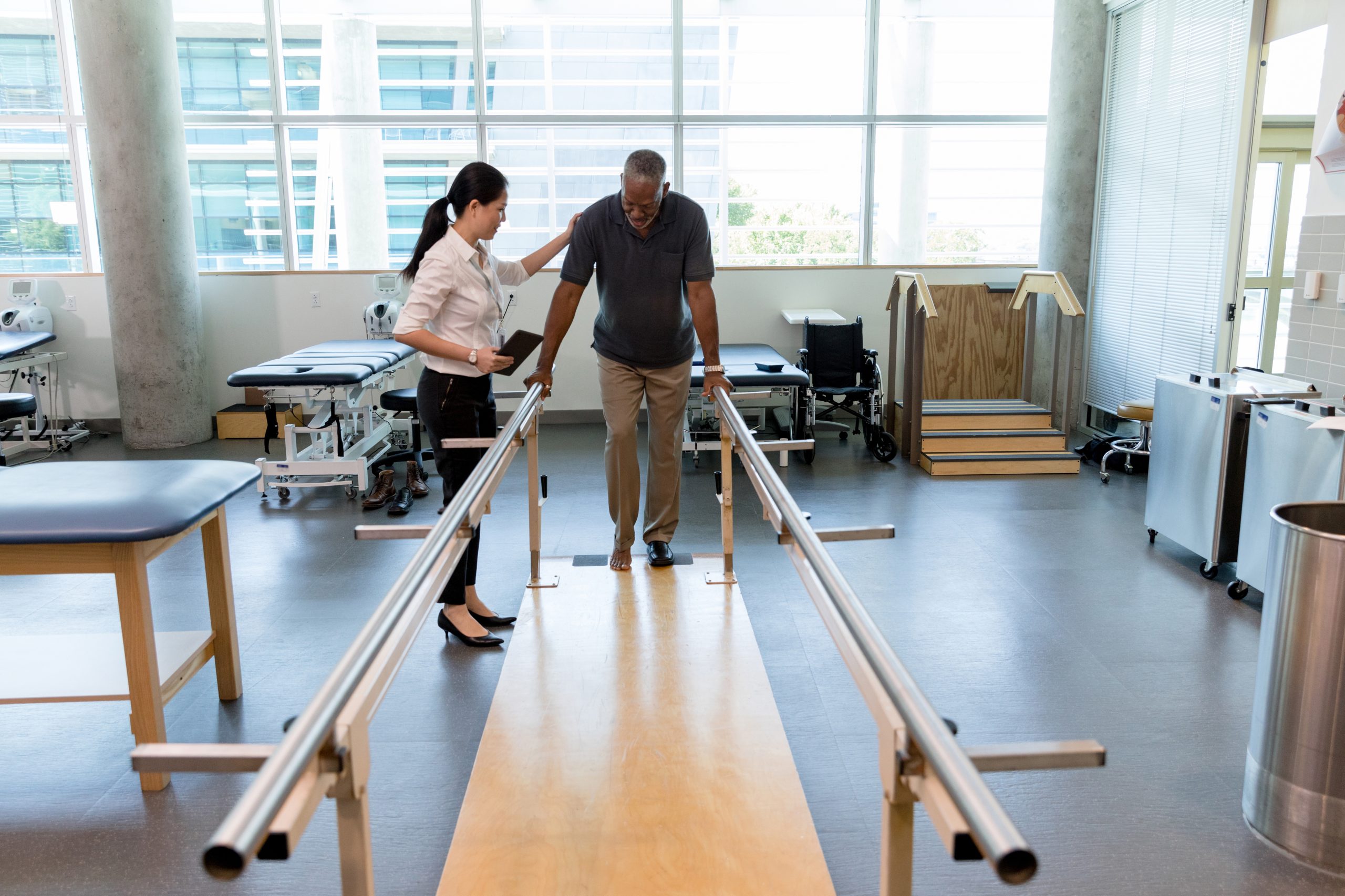 Female physical therapist helps a senior man walk following a stroke. The man is using parallel bars in a rehab center.